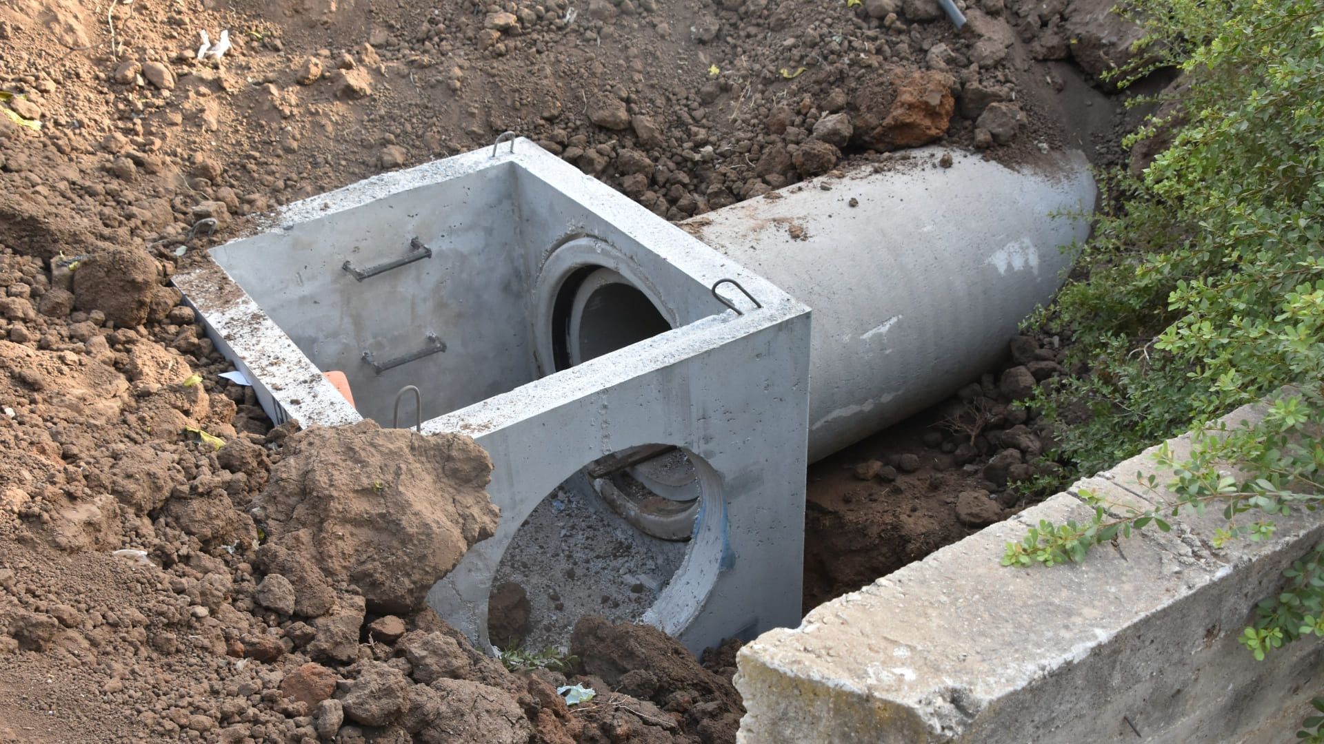 Concrete pipe and square drain in a dirt trench next to a stone curb and greenery.