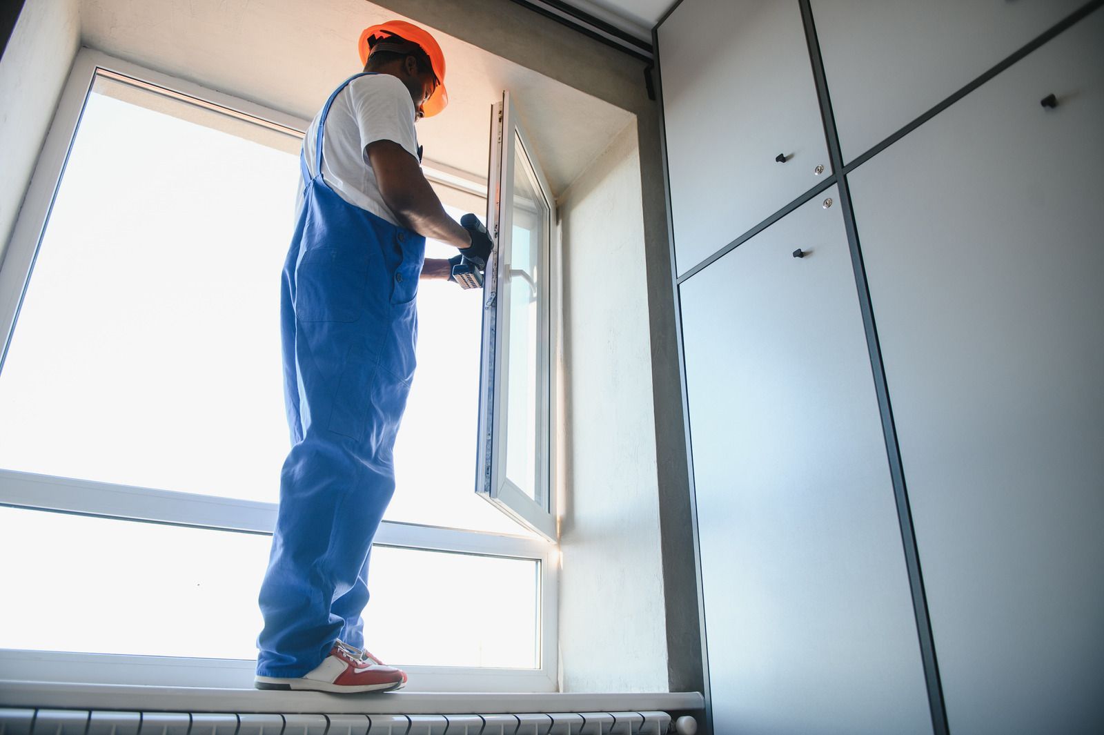 Construction worker in blue overalls and hardhat installing a window.