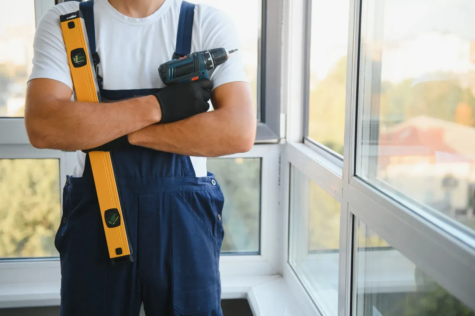 Worker in blue overalls holding a level and drill near a window.