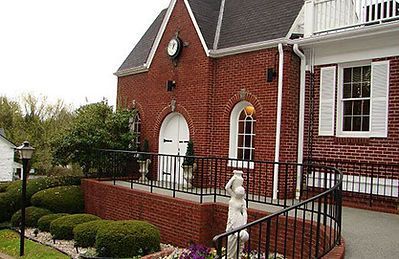 A red brick building with an arched entrance, a clock on the gable, a wheelchair ramp, and a white statue in the foreground.