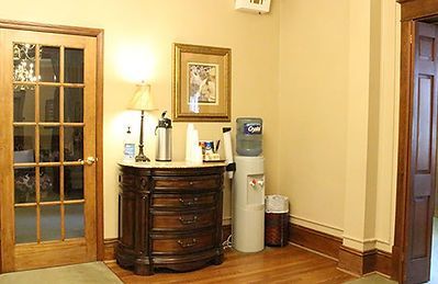 A wooden chest with a lamp and water dispenser in a hallway with two wooden doors and a small wastebasket.