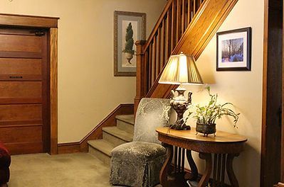 A cozy indoor hallway featuring a wooden staircase, a patterned armchair, and a lamp on a small side table.