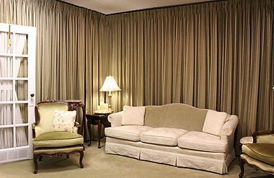 A cozy living room with a neutral-toned sofa, two armchairs, a side table with a lamp, and floor-to-ceiling beige curtains.