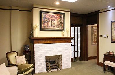 A white brick fireplace with a wooden mantel, topped by a framed painting, in a room with a green chair and dark woodwork.