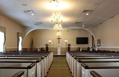 Interior view of a chapel with rows of wooden pews facing a central altar featuring a cross and two floor lamps.