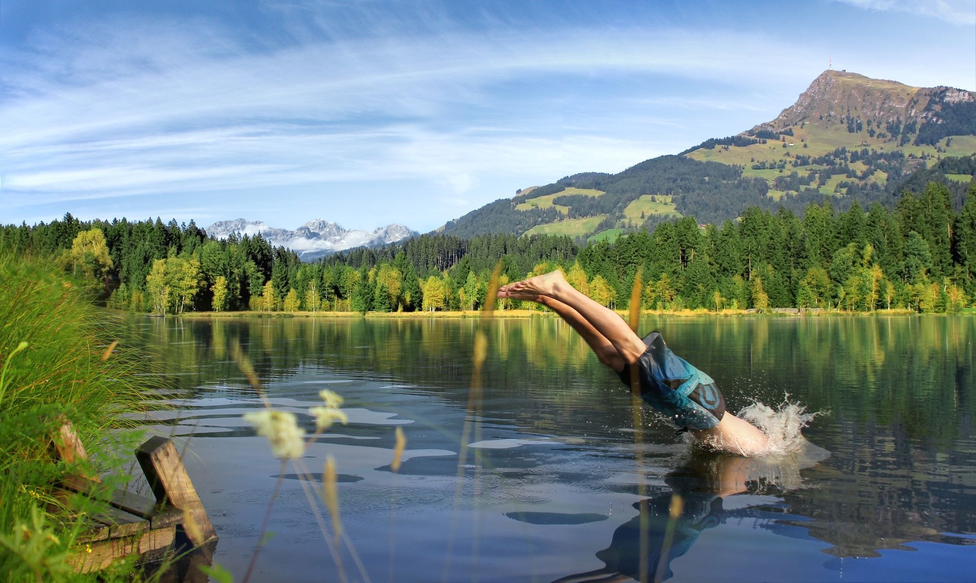 Eine Person taucht in einen See vor der Kulisse der Berge. Grüne Bäume umgeben das Wasser unter einem blauen Himmel.