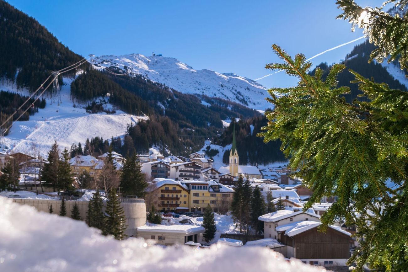 Schneebedecktes Bergdorf in einem Tal mit Skiliften und schneebedeckten Hängen, klarem blauen Himmel und Kiefern.