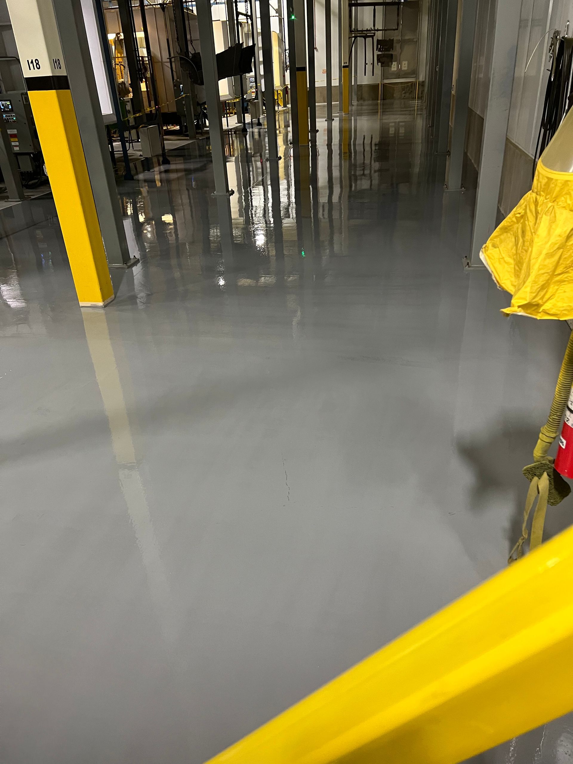 Gray-floored industrial interior reflecting overhead lights. Yellow safety poles and a railing are in the foreground.