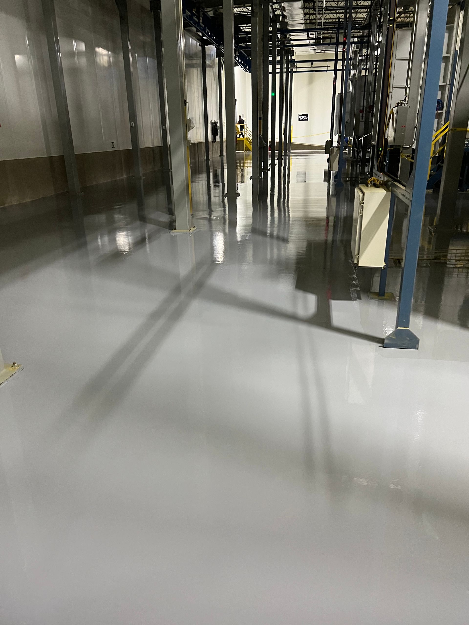 Shiny gray industrial floor in a warehouse-like setting. Reflective surface, columns, and overhead piping visible.