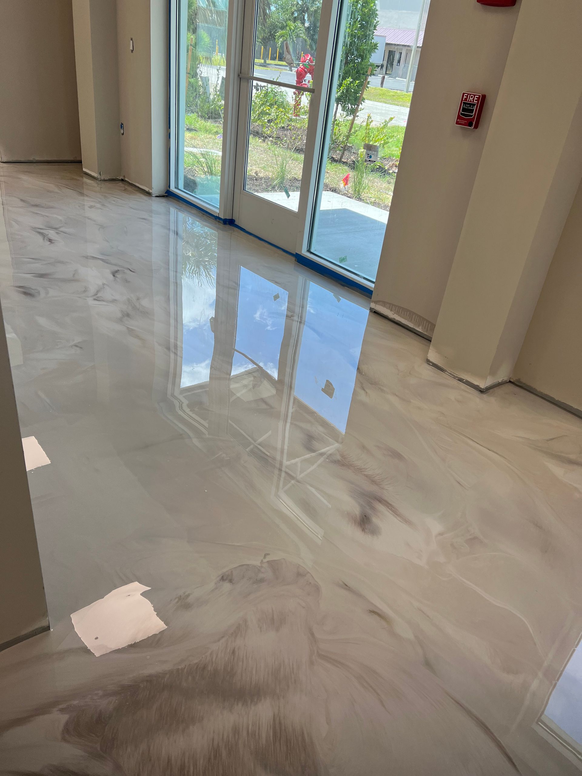 Shiny, light-colored epoxy flooring in a building. Glass doors with reflections. Beige walls and a fire alarm.