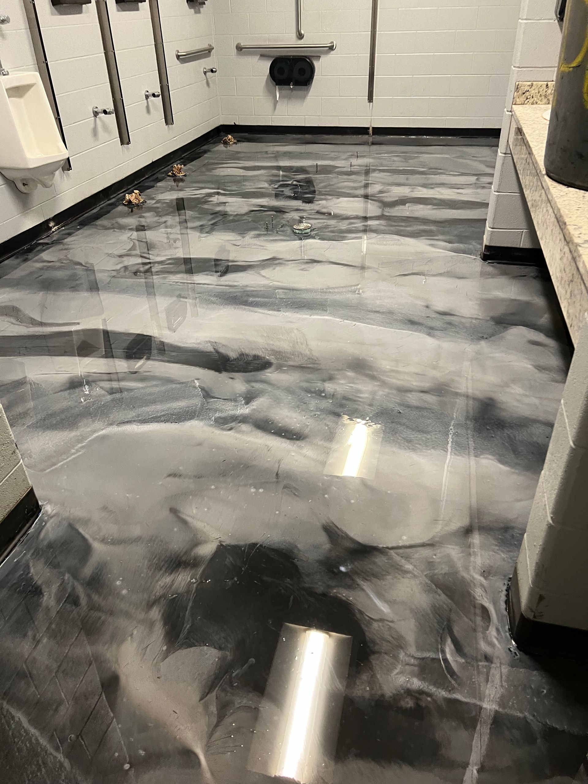 Bathroom with metallic epoxy floor, debris scattered. White walls, urinals visible.