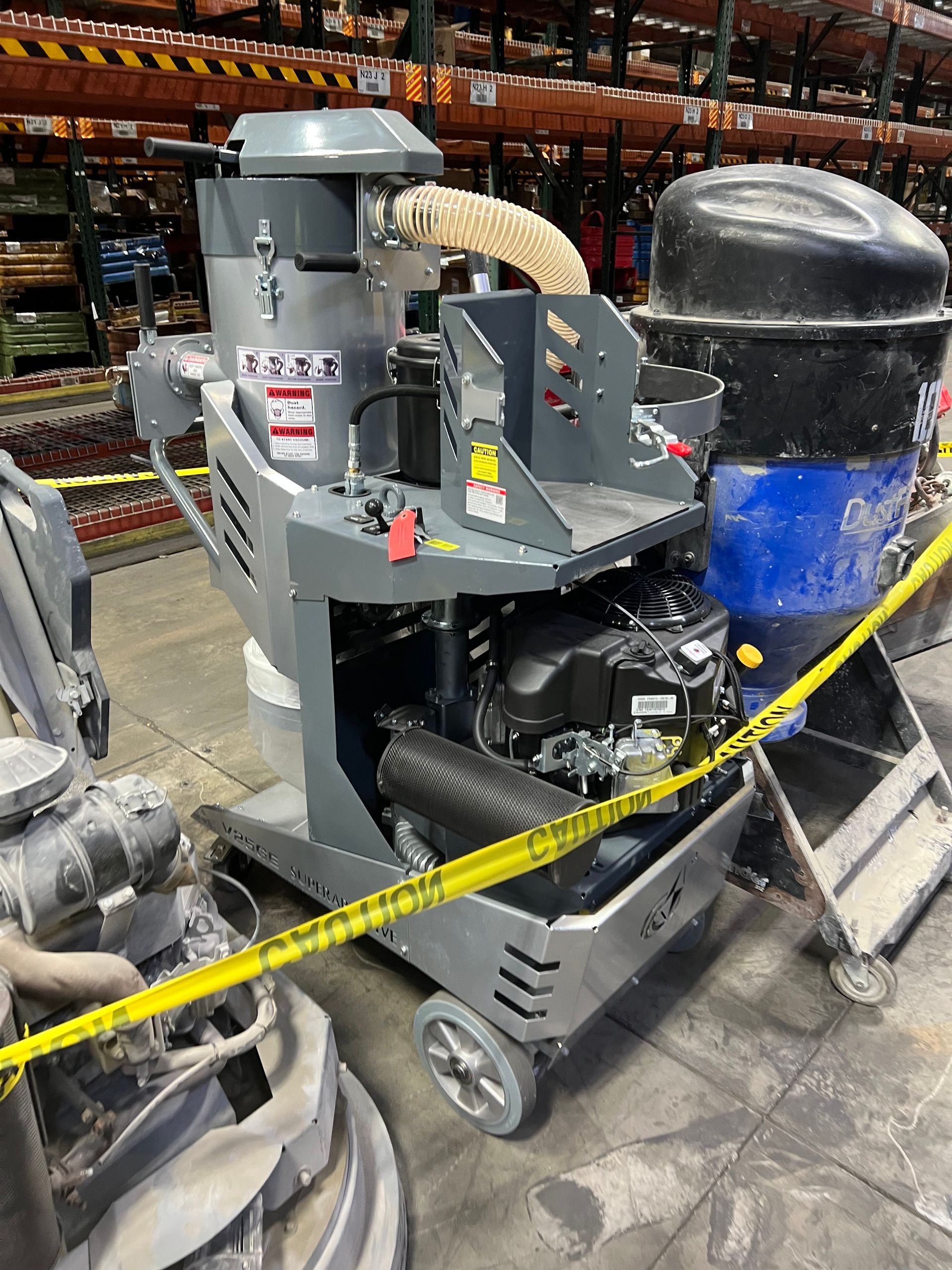 Gray and black industrial machine with a hose and a large blue container, yellow caution tape in a warehouse.