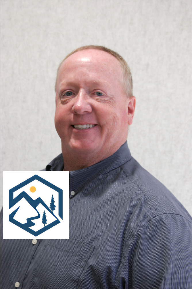 A man in a blue shirt is smiling with a mountain logo behind him