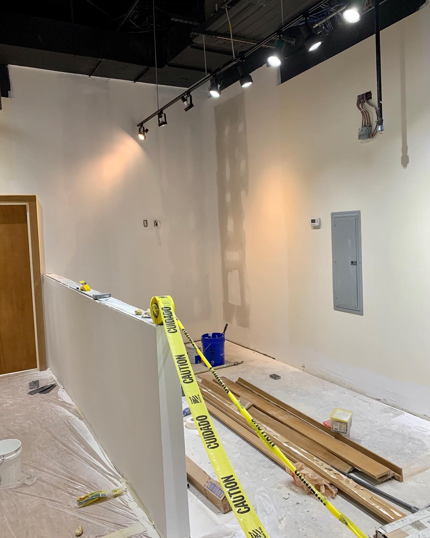 An interior room under construction with white walls, track lighting, a partition with yellow caution tape, and floor debris.