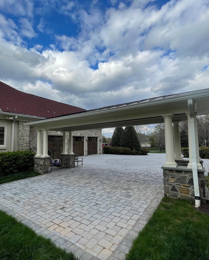 A stone-paved driveway leads to a three-car garage under a white pergola with stone pillars against a cloudy sky.