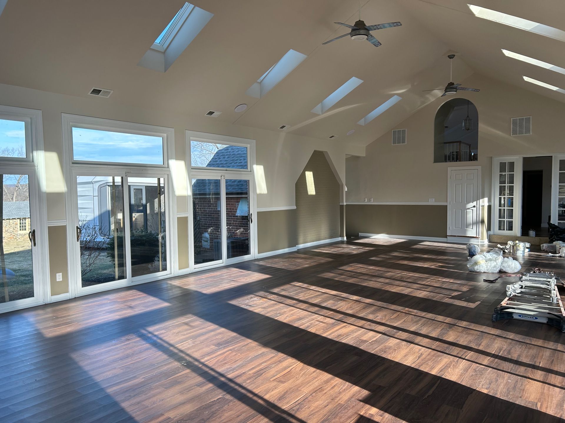 A spacious, sunlit room with high ceilings, skylights, dark wood flooring, and large windows leading to a patio.