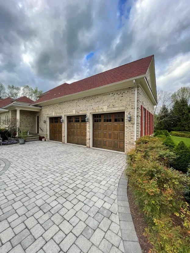 A three-car garage with a red tile roof and light stone exterior, fronted by a patterned paver driveway under a cloudy sky.