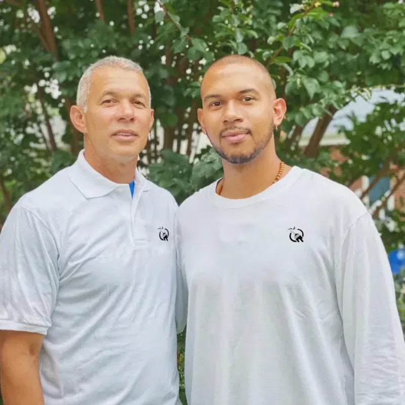 Two men stand side-by-side in front of a tree, wearing matching white shirts with a small circular logo on the chest.