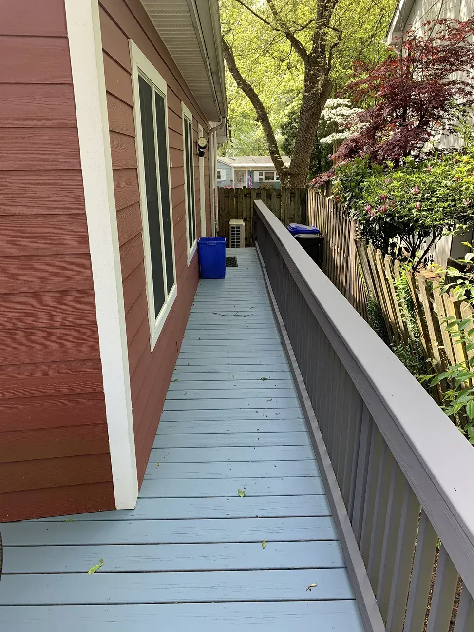 A narrow outdoor wooden deck path painted light blue, running alongside a red house wall with a wooden railing on the right.