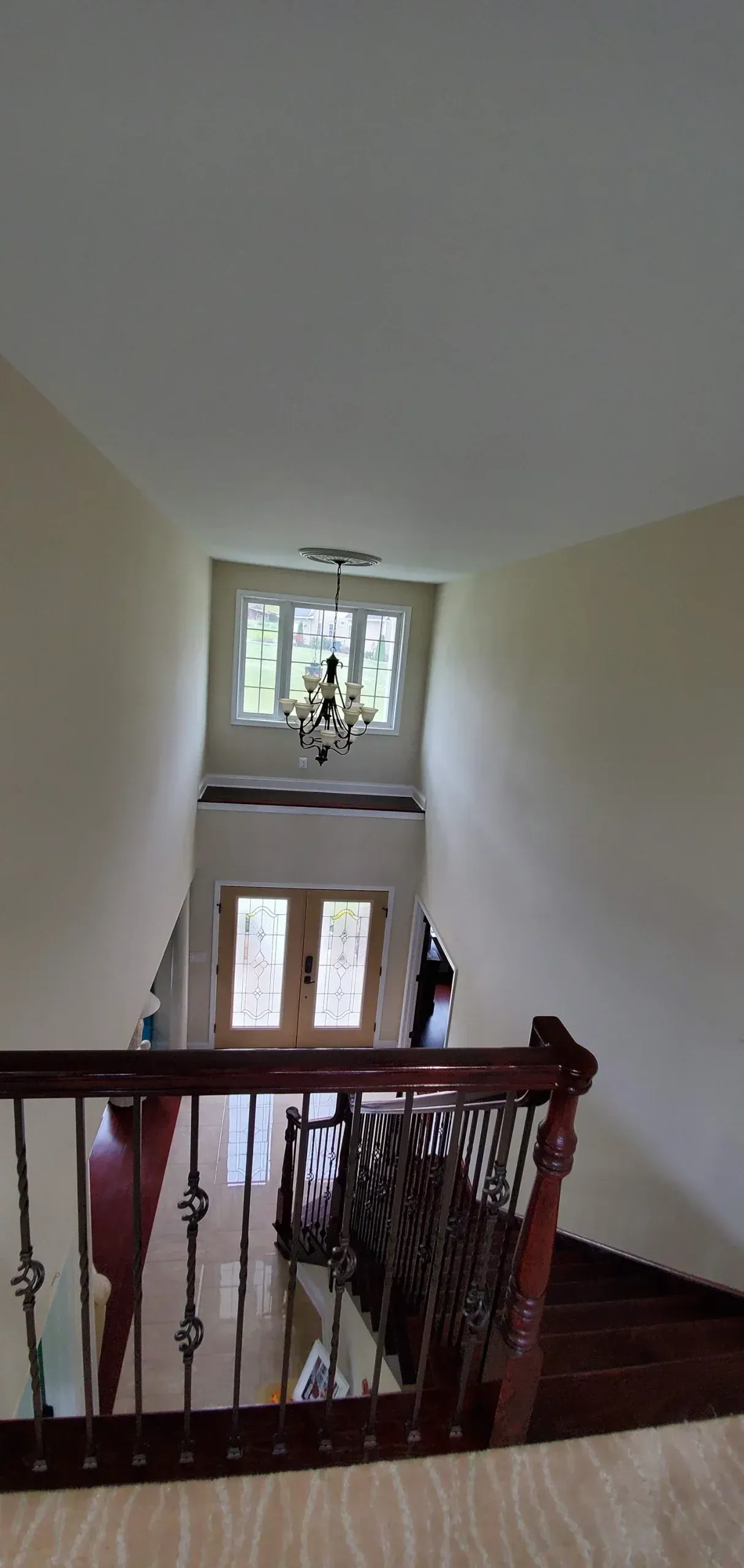 A high-angle view looking down a wooden staircase with decorative iron railings toward a front entryway and chandelier.