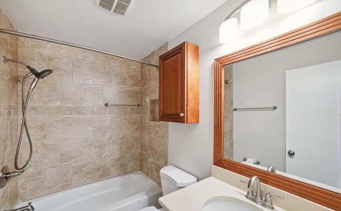 A brightly lit bathroom with a tan tiled shower, a white bathtub, a wood cabinet, and a mirror above a vanity sink.