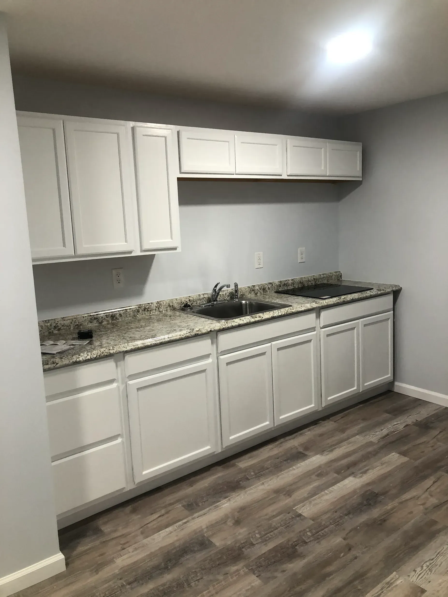 A kitchen area with white cabinets, granite countertops, a sink, and dark wood-look vinyl flooring against gray walls.