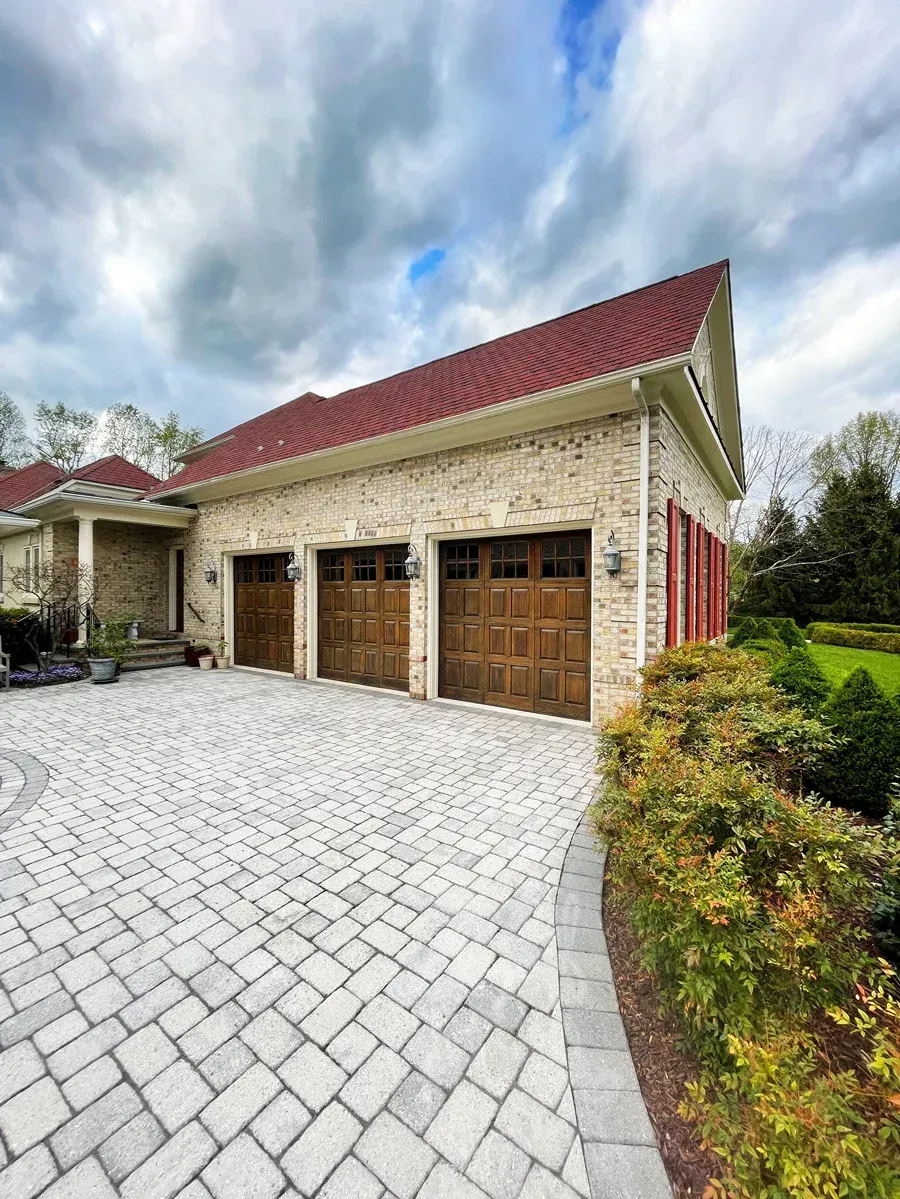 A tan brick garage with three wooden doors, a red tiled roof, and a patterned stone driveway under a cloudy sky.