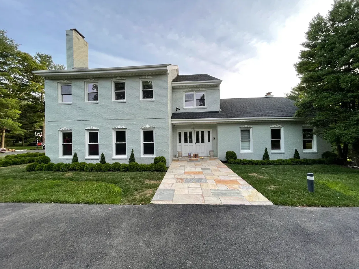 A light blue, two-story colonial-style house with a stone walkway leading to the front door, surrounded by a green lawn.