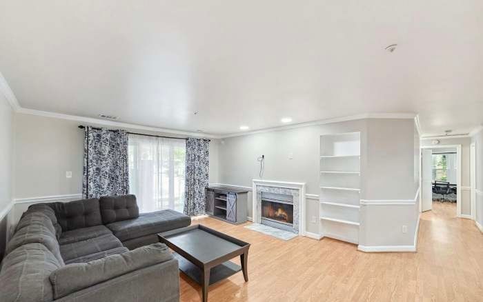 A light-colored living room with a large gray sectional sofa, a coffee table, a fireplace, and built-in shelving.