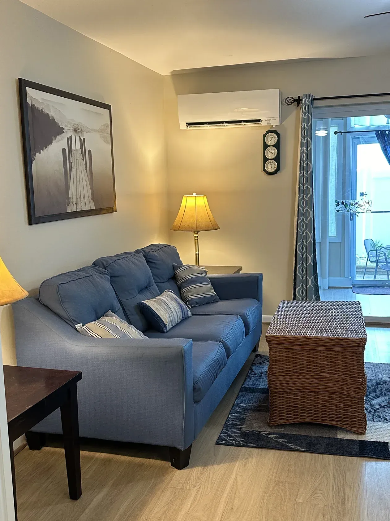 A blue sofa with patterned pillows, a wicker chest, a lamp, and wall art in a room with a patio door and AC unit.