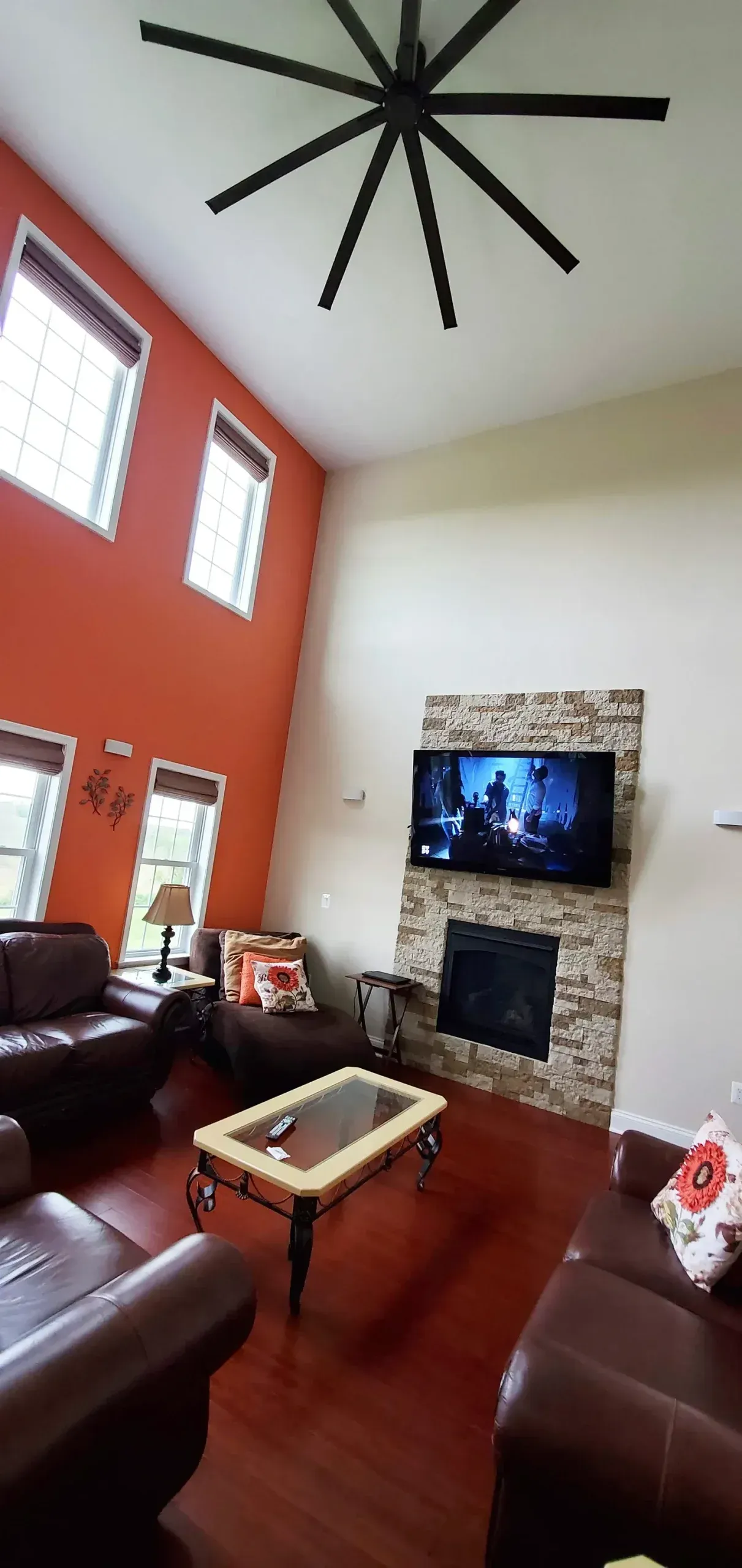 A tall living room with orange walls, a large ceiling fan, a stone fireplace, dark leather couches, and a coffee table.