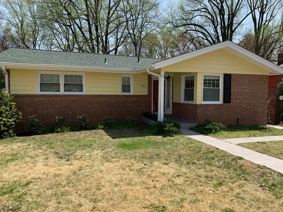 A single-story house with yellow siding and red brick exterior, a green shingled roof, and a paved walkway in front.
