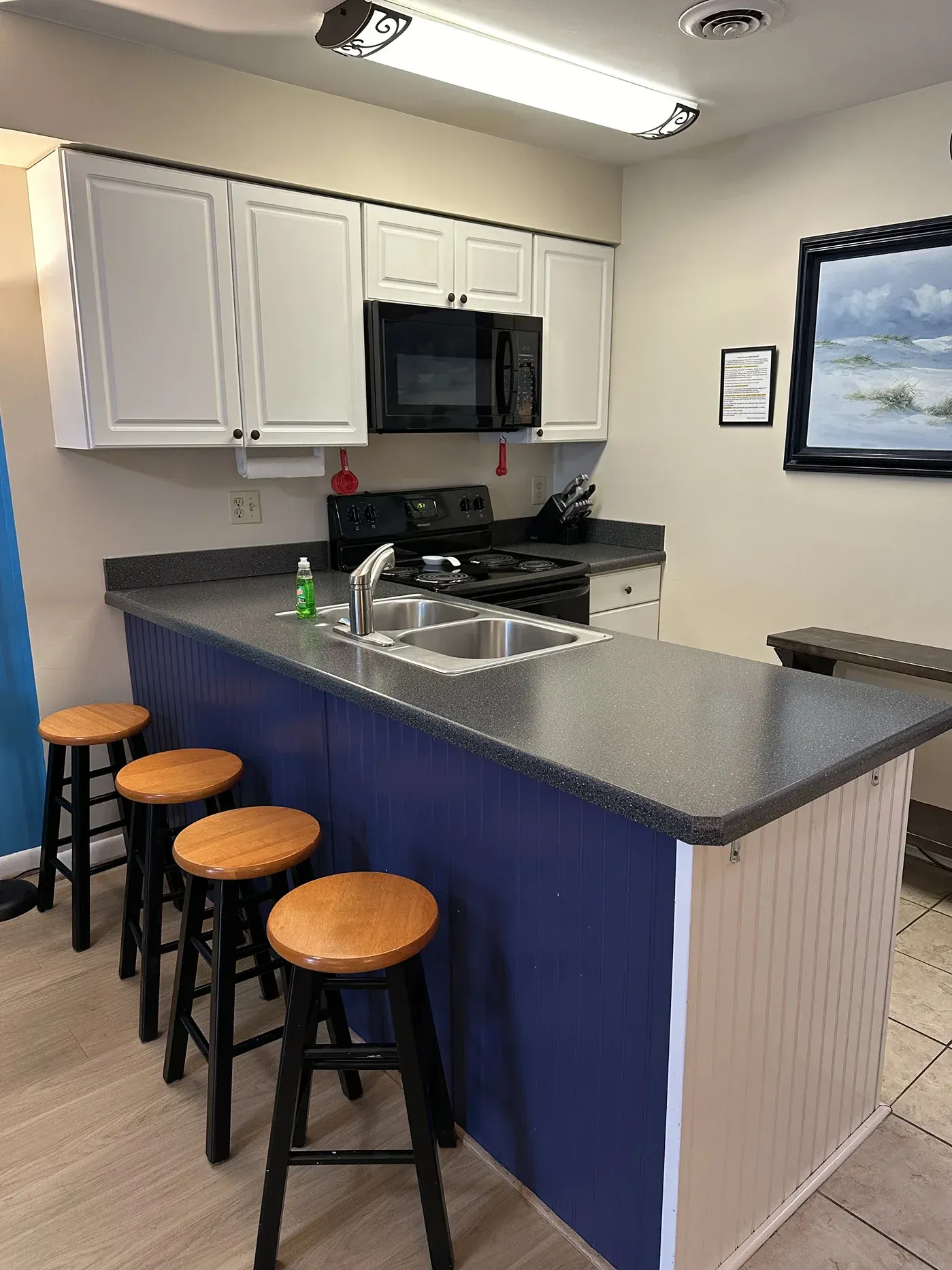A kitchen island with four bar stools in front of a dark counter, white cabinets, and a microwave in a brightly lit room.