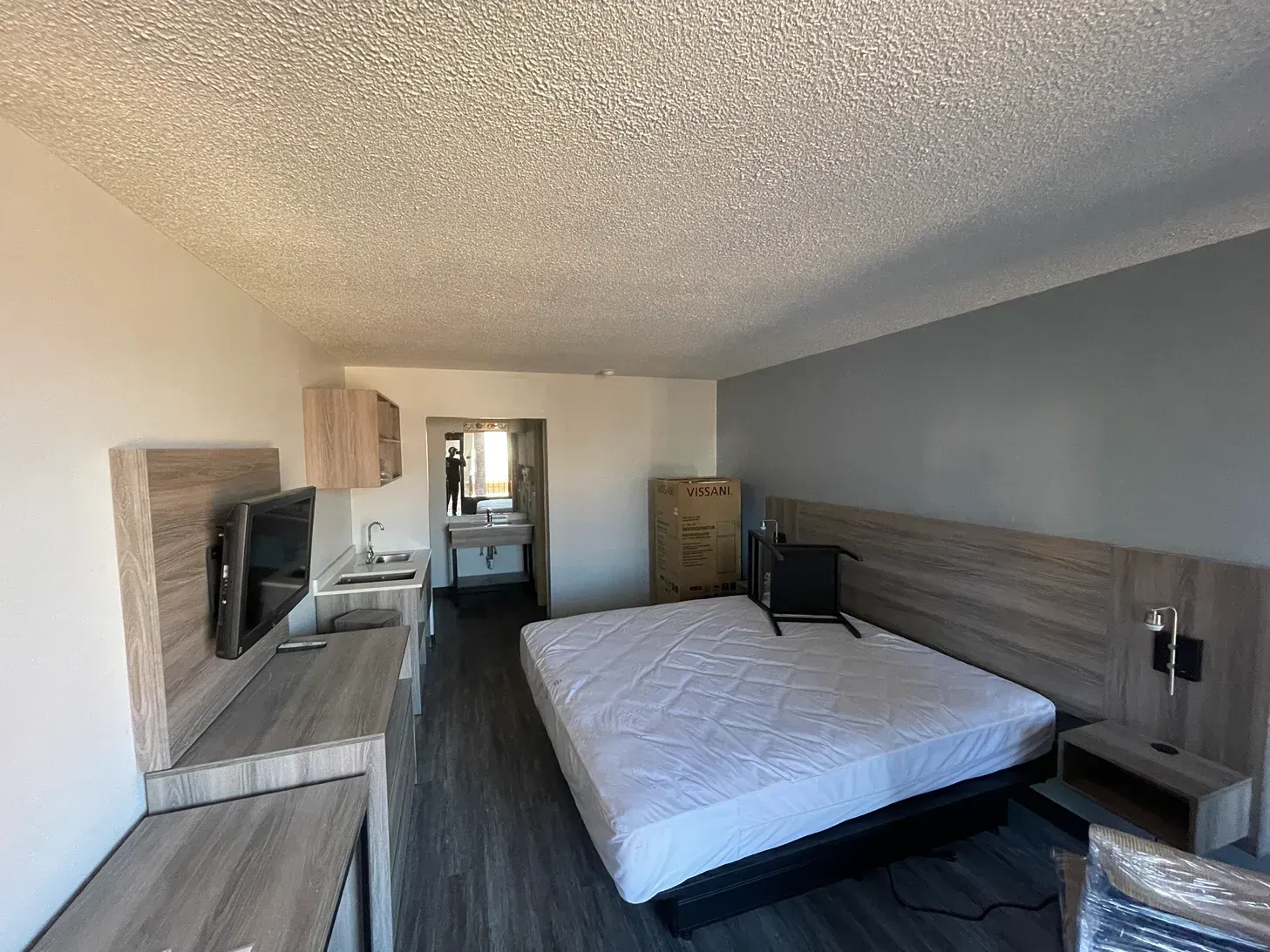 A modern hotel room with a large white bed, a grey wall, a flat-screen TV on a wooden stand, and a bathroom entrance.