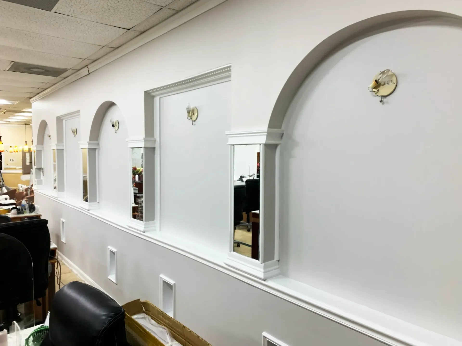 A row of white, arched wall alcoves, separated by mirrored pillars, along a salon wall with chairs in the foreground.