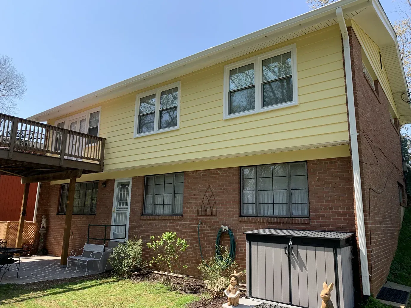 A two-story house with yellow horizontal siding on the top floor and brick on the bottom, featuring a deck and patio.
