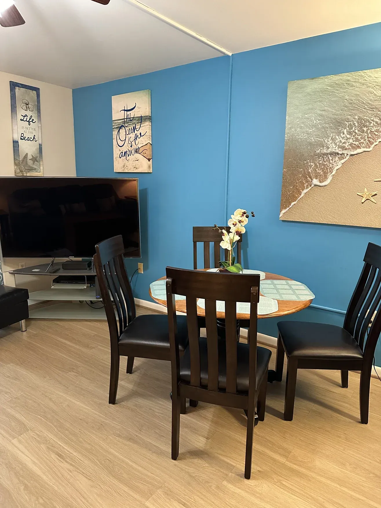 A dining table with four dark chairs sits in a room with a blue accent wall, a television, and beach-themed wall art.
