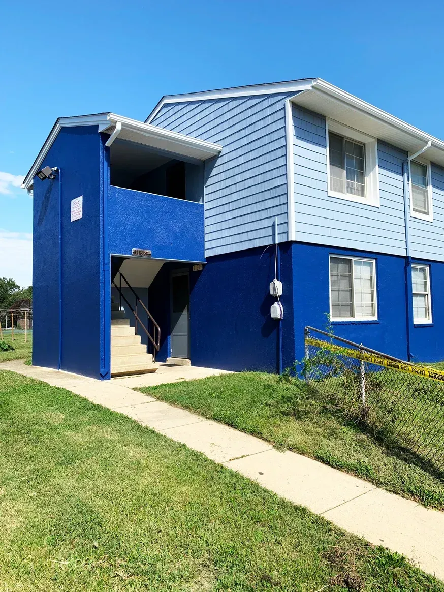 A two-story apartment building with blue and light blue siding, a central staircase, and yellow caution tape on the lawn.