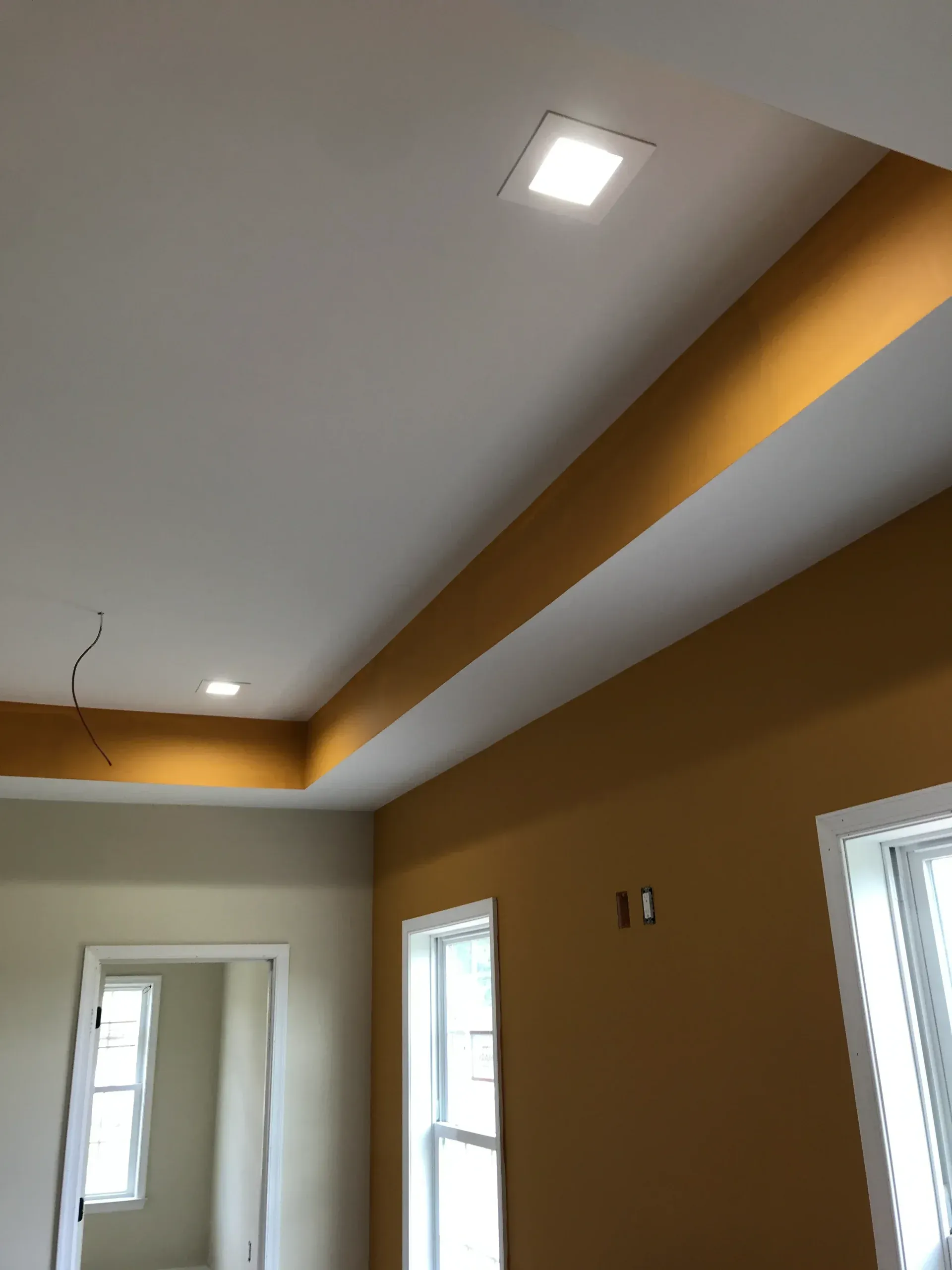 An interior view showing a white tray ceiling with recessed square lights, mustard yellow walls, and white trim windows.