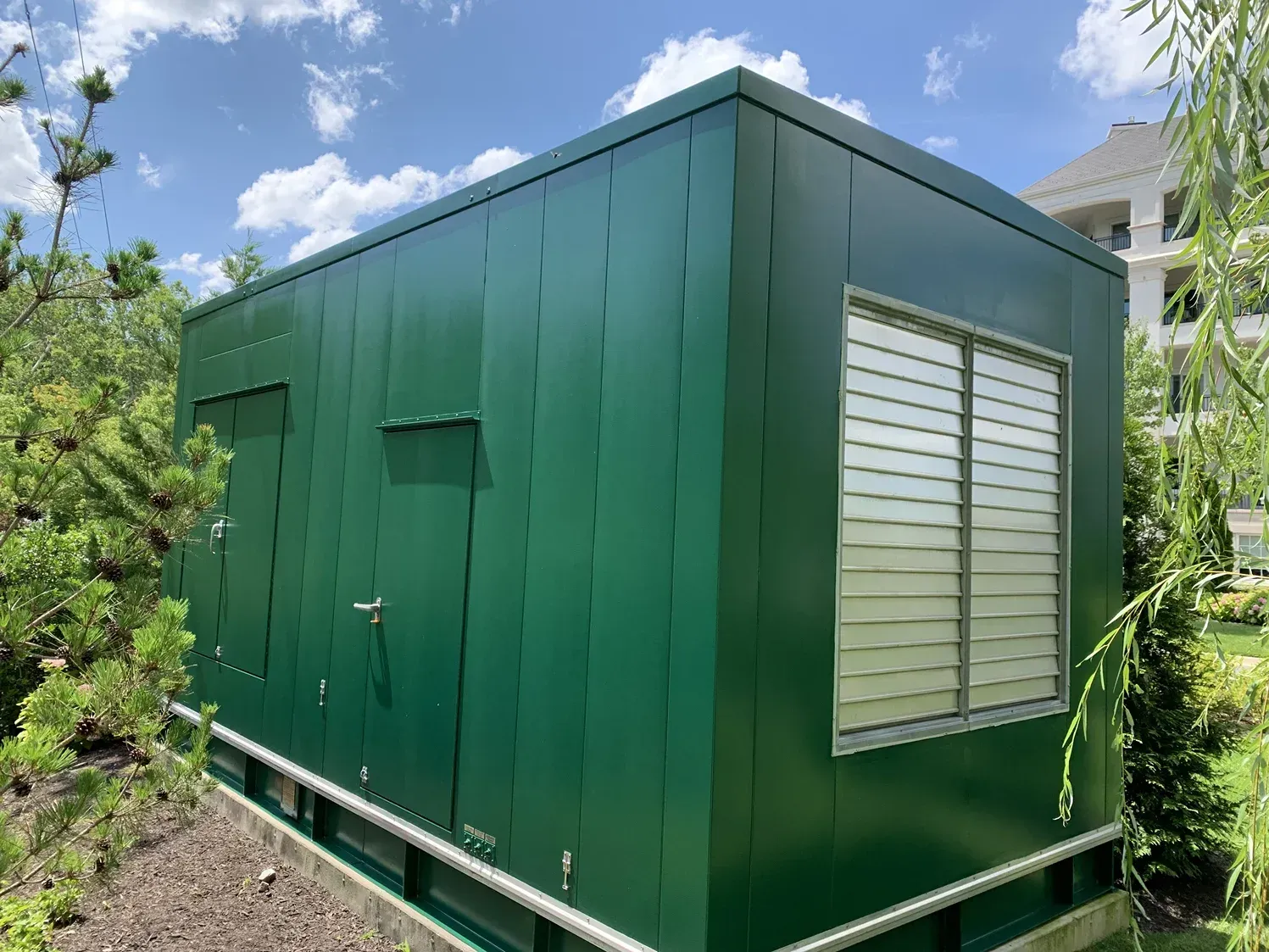 A dark green, rectangular utility enclosure with a louvered vent and service doors, located in a landscaped area.
