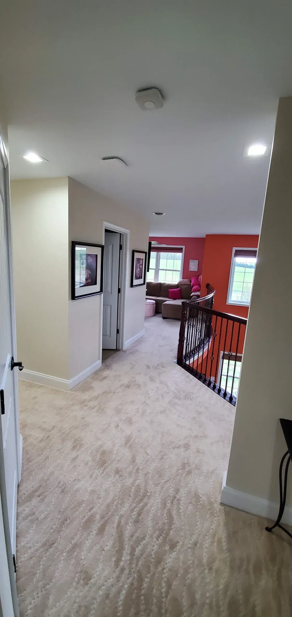 A hallway with beige carpeting leads toward a landing with dark wood railings and an orange-walled room in the background.