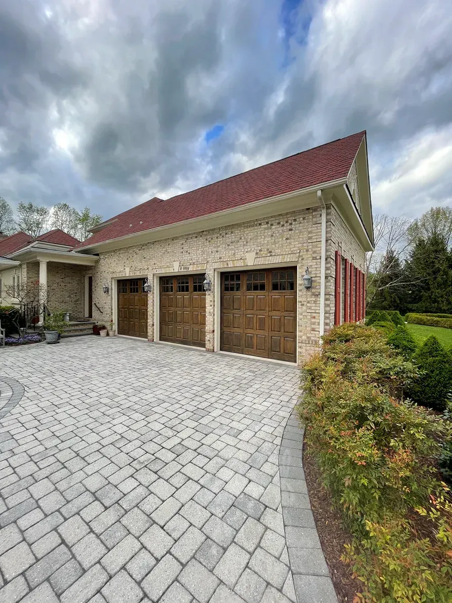 A three-car garage with wooden doors and a red tiled roof, featuring a textured stone facade and a paved driveway.