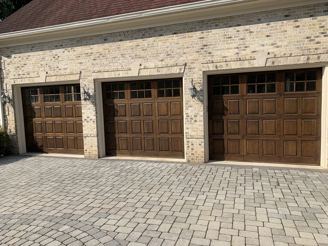 Three dark wood garage doors with window panels set in a light stone building, with a gray cobblestone driveway.