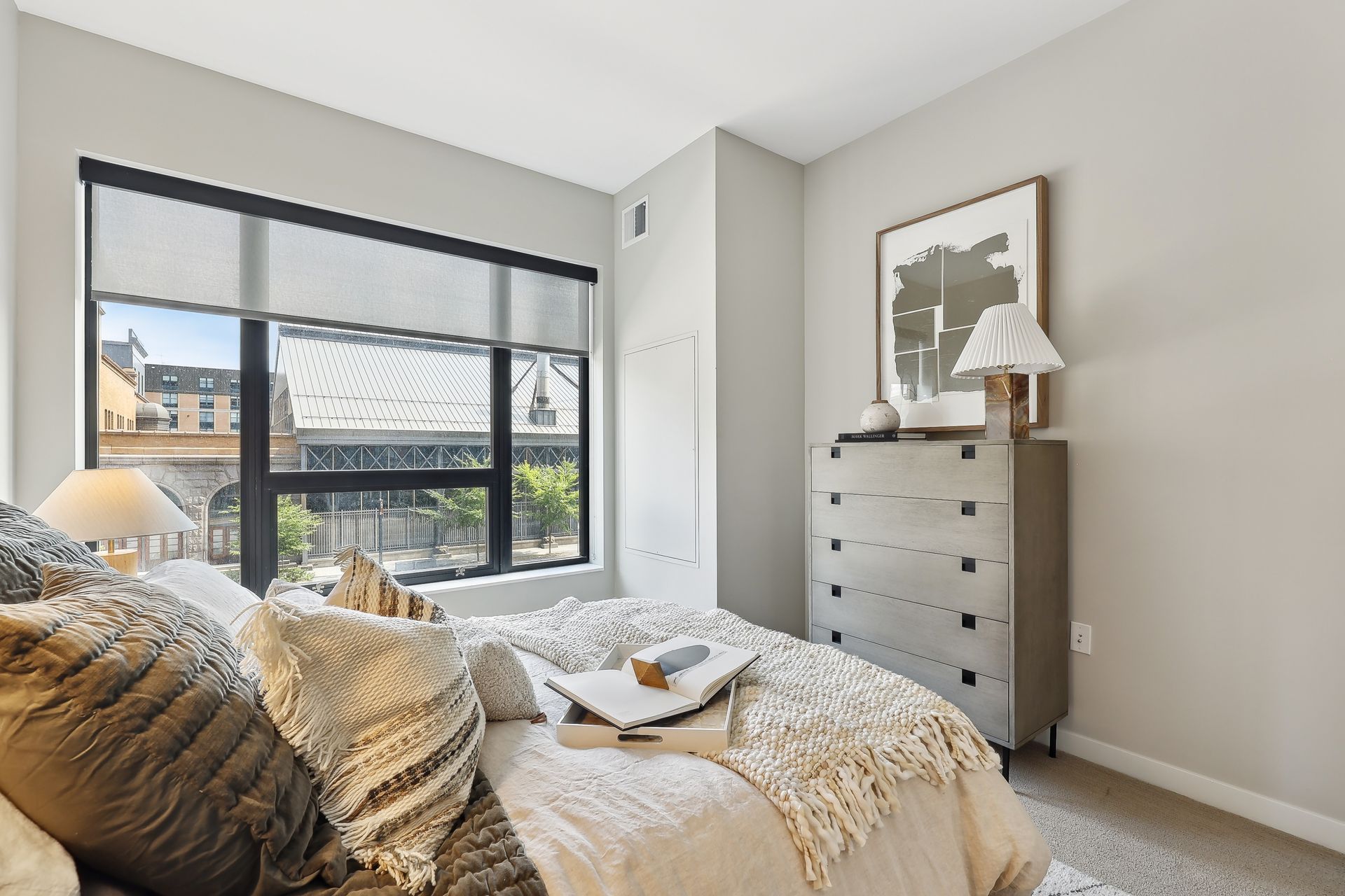 Bedroom with bed, nightstand, art, and large window with roller shade. Neutral colors.