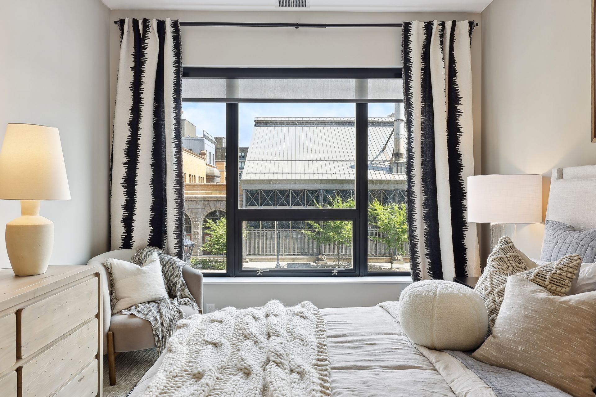 Bedroom with bed, window, curtains, a dresser, and a chair. Beige walls and decor, daylight.