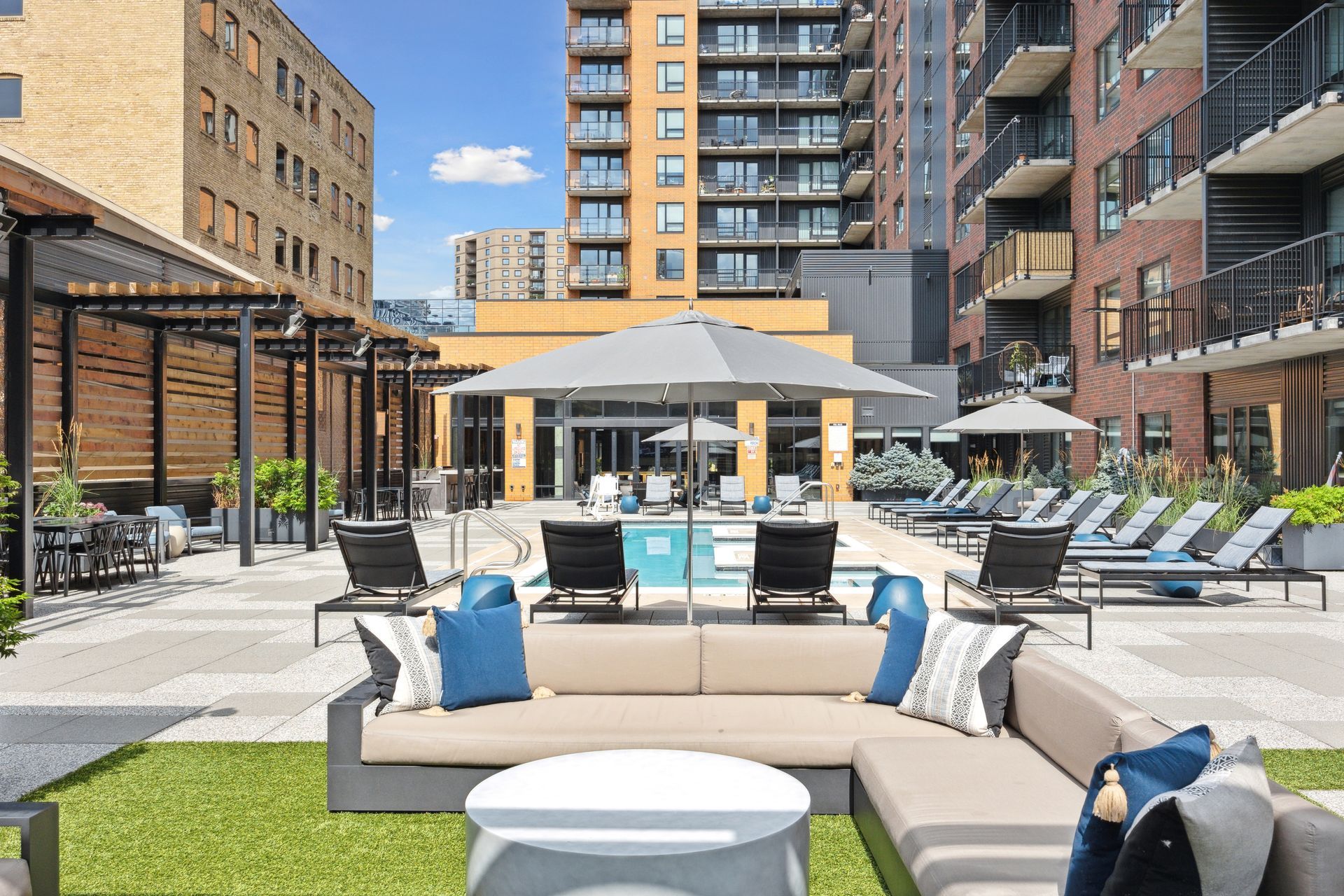 Outdoor lounge area with pool and modern high-rise buildings. Comfortable seating with blue pillows, umbrellas.