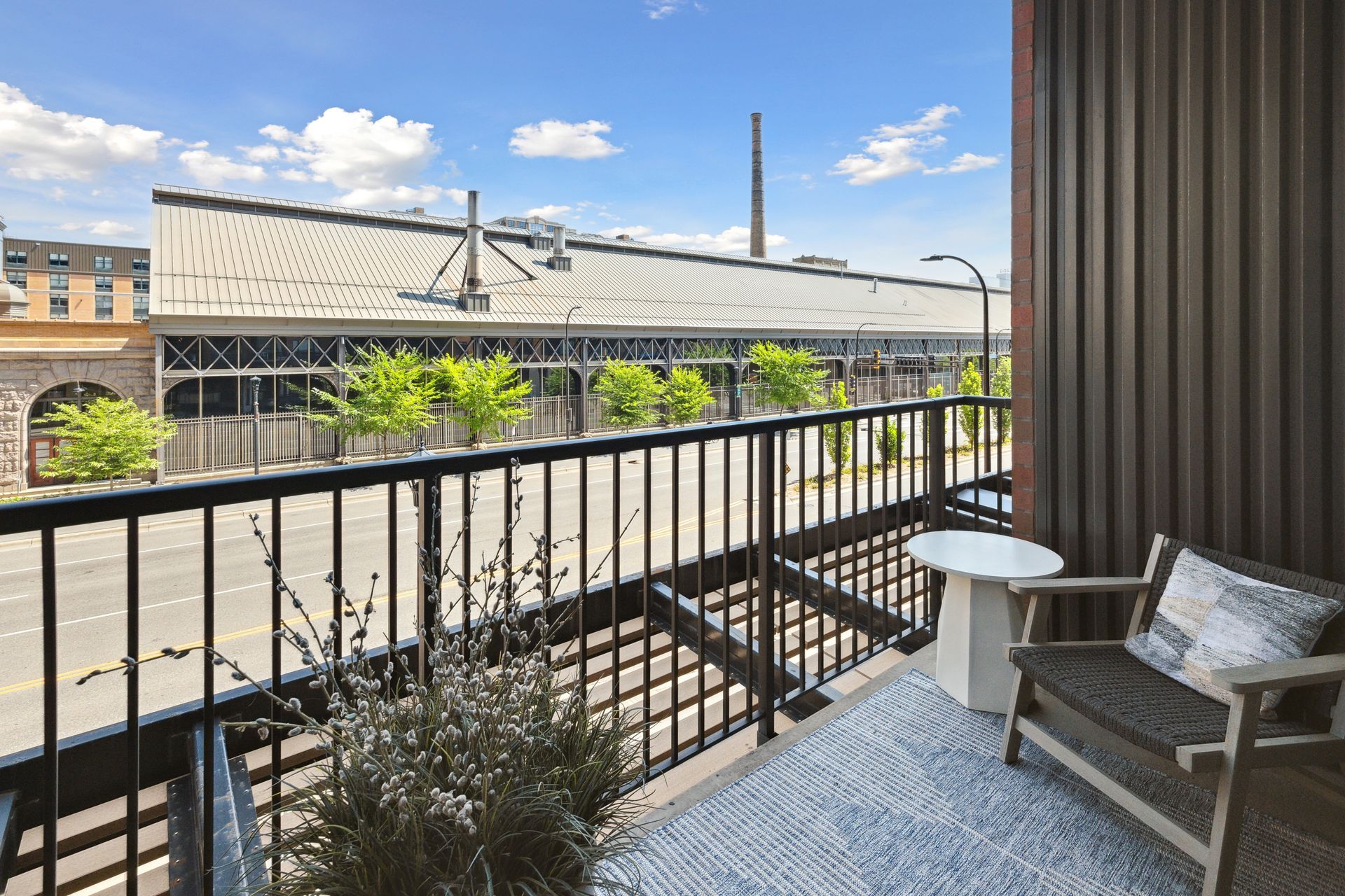 Balcony view of street and large industrial building with greenery. Chair, table, plants visible.