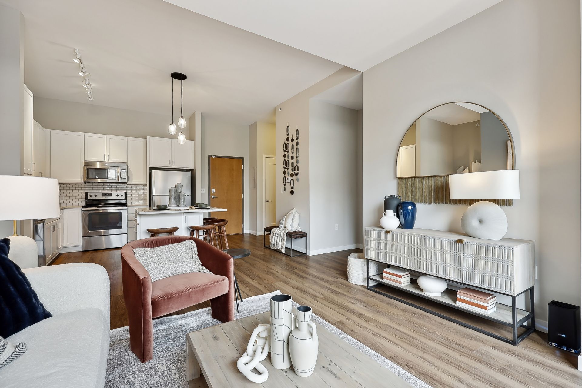 Living room with open kitchen: white walls, light wood floors, modern furniture, and large arched mirror.