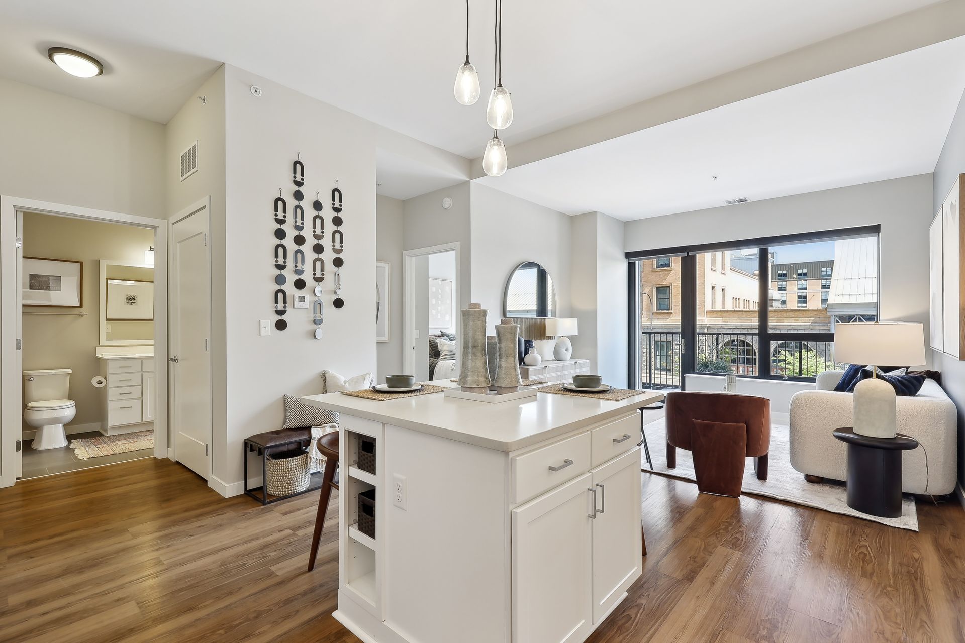 Modern apartment interior with open kitchen, living area, and bathroom visible. White walls, hardwood floors, and bright lighting.