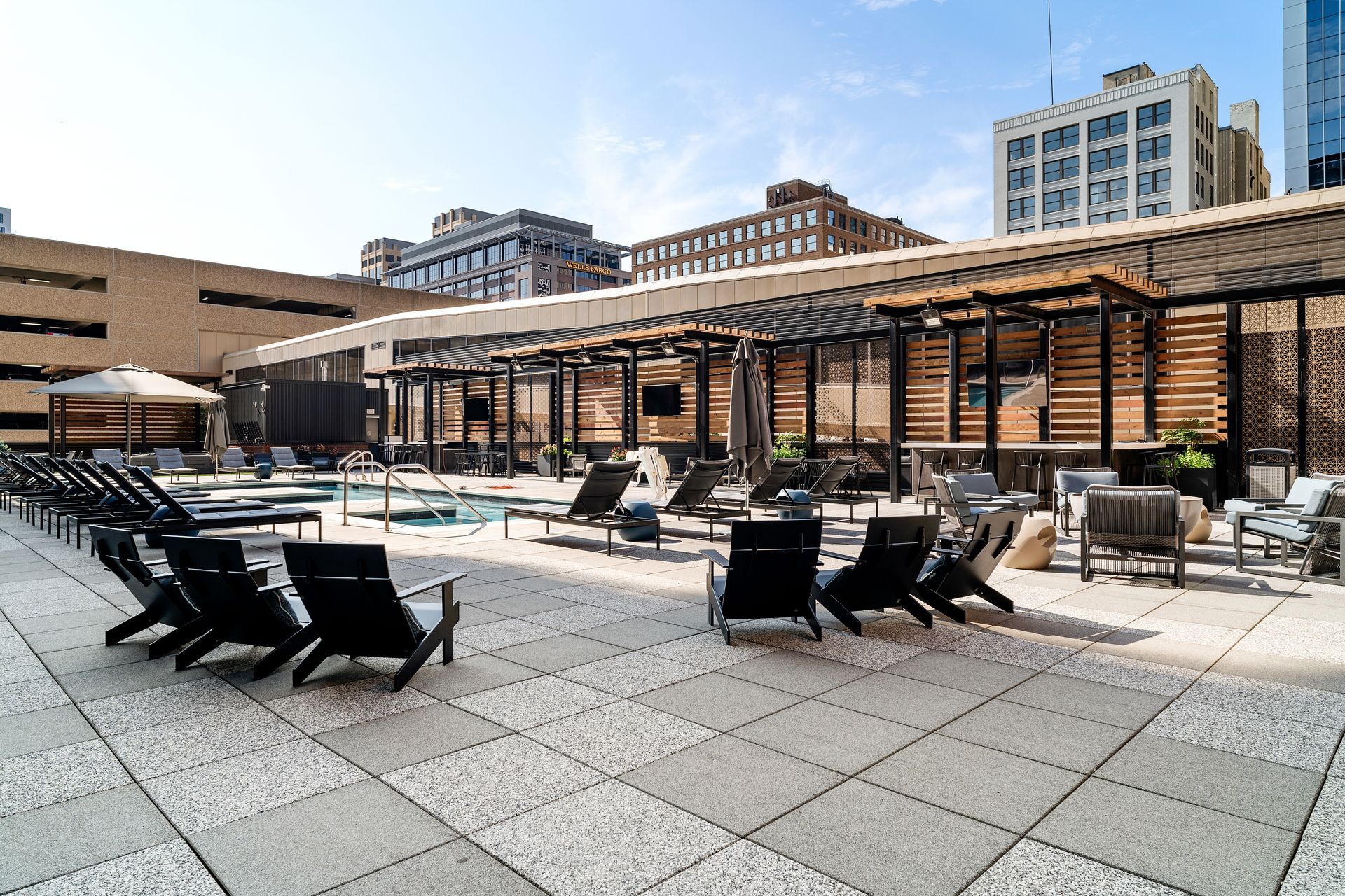 Rooftop pool deck with lounge chairs, pergola, and city buildings in the background. Sunny day.